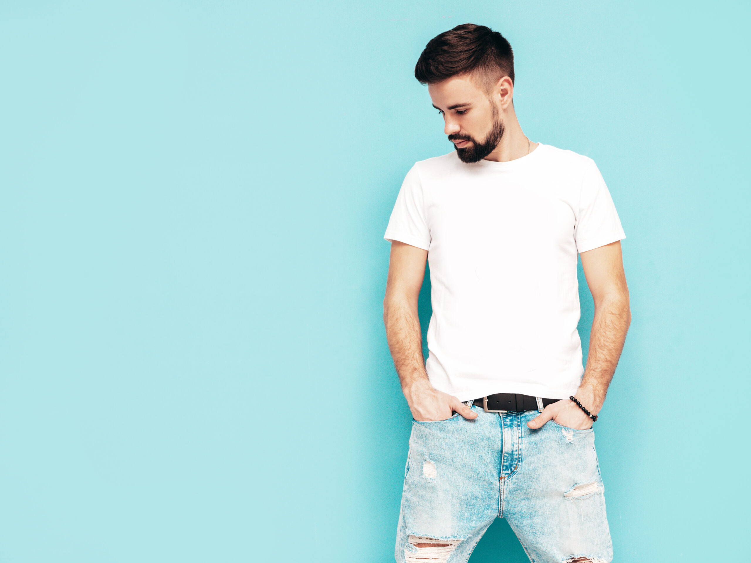 Portrait of handsome confident stylish hipster lambersexual model.Man dressed in white T-shirt and jeans. Fashion male isolated on blue wall in studio