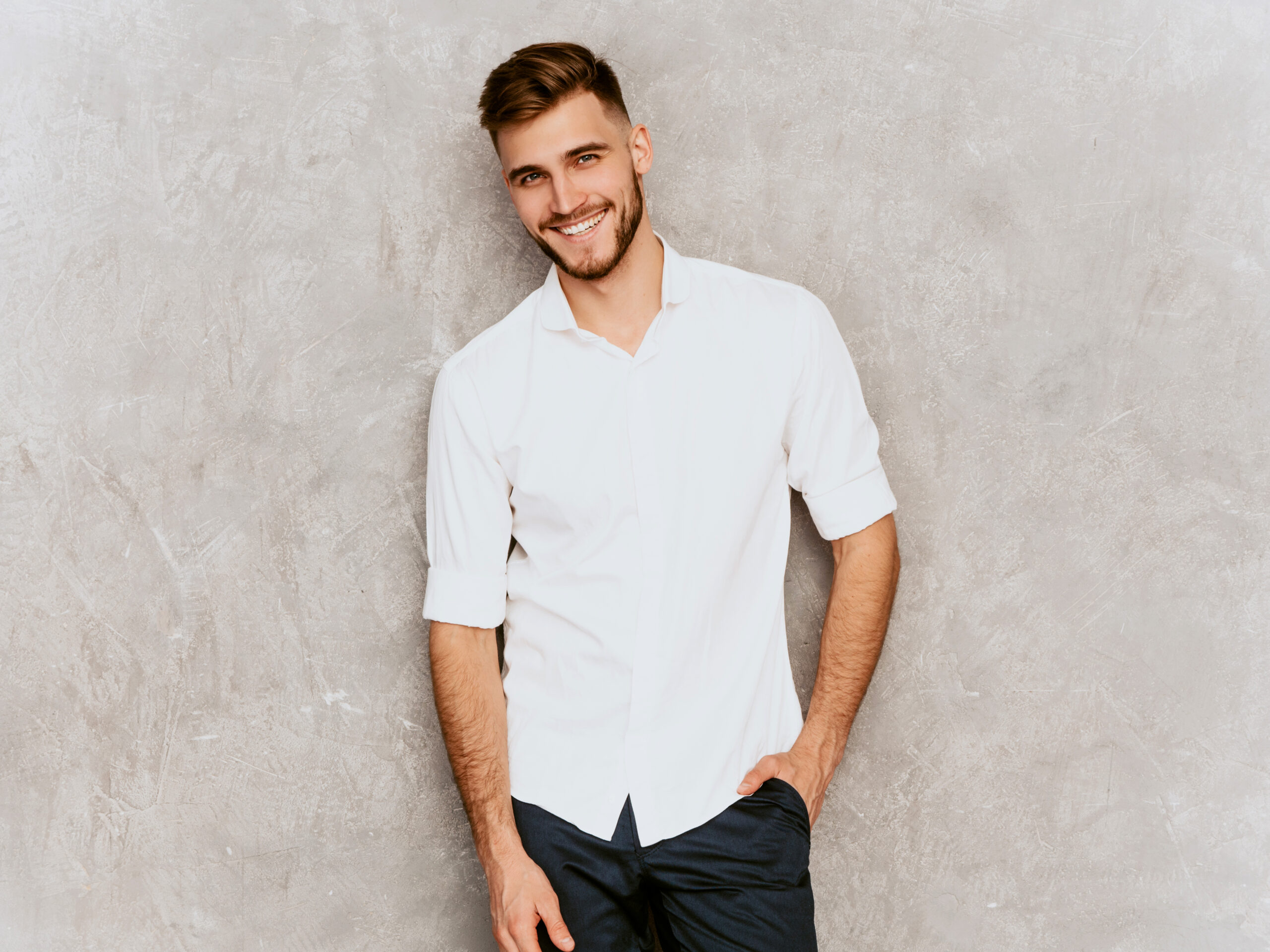 Portrait of handsome smiling hipster lumbersexual businessman model wearing casual summer white shirt. Fashion stylish man posing against gray wall
